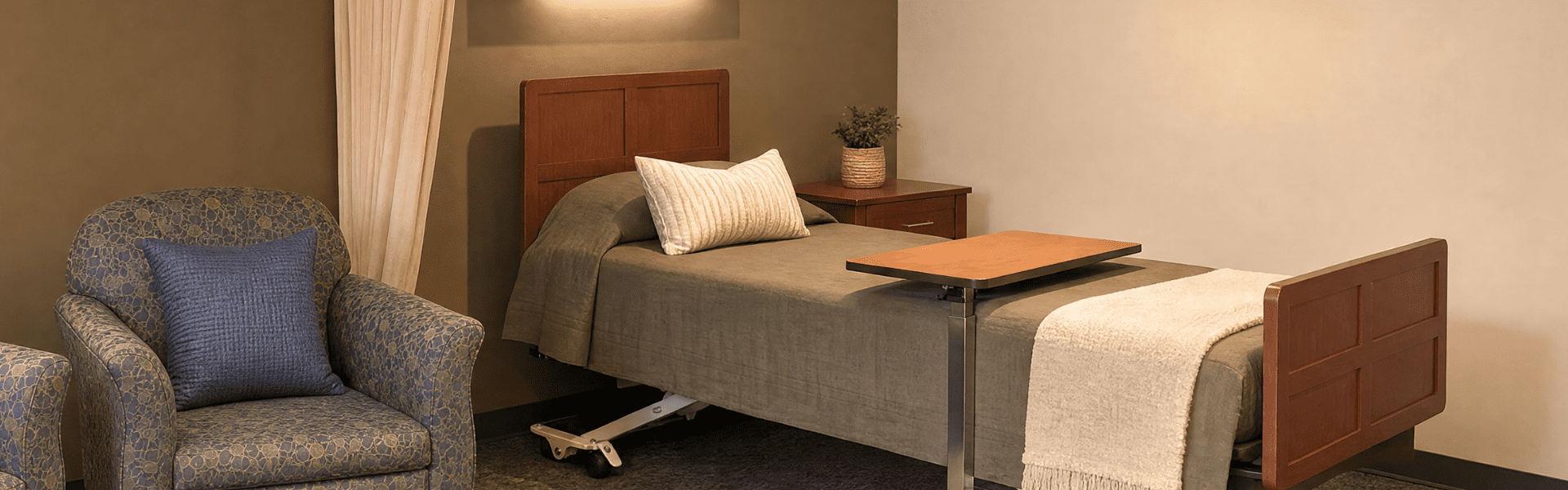 A hospital or care facility room with a bed that has a wooden headboard and footboard, a gray bedspread, a beige blanket, and a pillow. There is a bedside table with a potted plant and a tray attached to the bed. An upholstered armchair with a patterned fabric and a dark blue pillow is next to the bed.