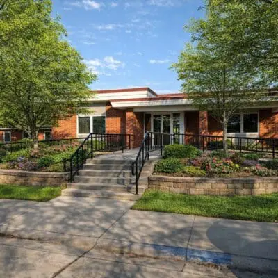 A single-story brick building with a white entrance door, surrounded by green trees and landscaped flower beds. A wheelchair ramp and steps lead up to the entrance, with a sidewalk in the foreground.
