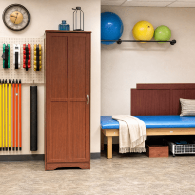 A room with a pegboard wall displaying colorful resistance bands and exercise mats, a tall wooden cabinet, a blue exercise bench with a gray pillow and white towel, and a shelf with blue, yellow, and green exercise balls.