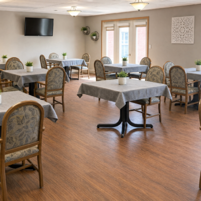 A dining room with multiple tables covered in gray tablecloths, each decorated with small potted plants. There are wooden chairs around the tables, large windows letting in natural light, a wall-mounted flat-screen TV, and light-colored walls with minimal decor.