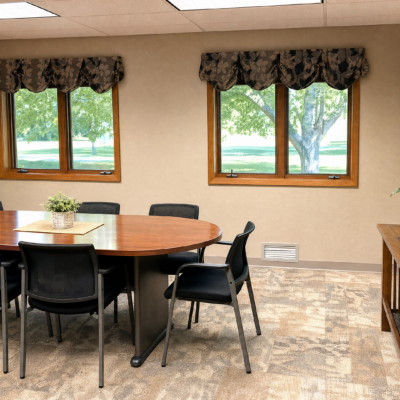 A round wooden table with a small potted plant in the center, surrounded by six black chairs, in a room with beige walls, two large windows with floral valances, and a view of green trees outside.