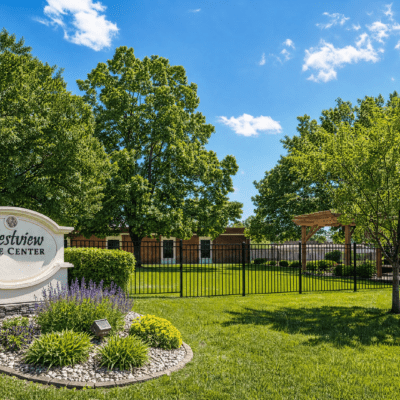 A well-maintained outdoor area with lush green grass, tall trees, and a clear blue sky with a few clouds. There is a fence and a building in the background, with a sign that reads 