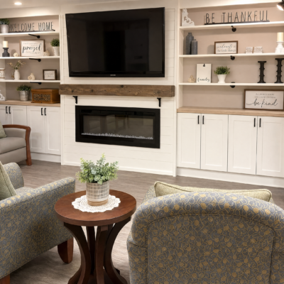 A cozy living room with a flat-screen TV mounted above a fireplace. White built-in shelves with decorative items and signs, including 