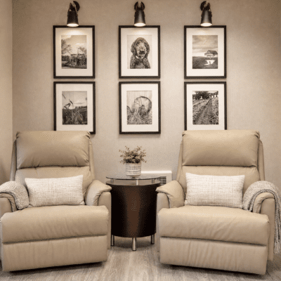 Two beige armchairs with pillows sit side by side, separated by a small round table with a potted plant. Behind the chairs, a wall displays six black-and-white framed photographs arranged in two columns. The lighting above highlights the pictures.