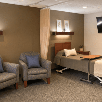 A bedroom with a bed, two gray armchairs with pillows, a bedside table with a plant, a wall-mounted shelf, and a wall-mounted TV.