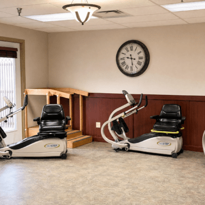 A room with two recumbent exercise bikes, one on each side, positioned on a carpeted floor. There is a wall clock showing 10:12, a wooden railing or platform in the background, a ceiling light fixture, and a window with vertical blinds on the left side.