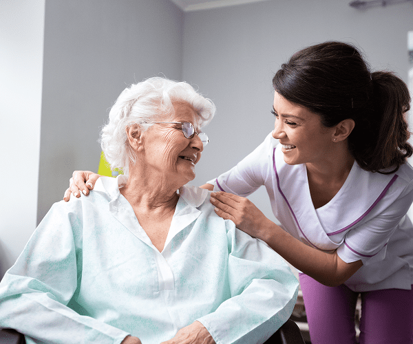 A healthcare worker smiling and talking to an elderly woman with white hair and glasses, who is sitting in a chair. The healthcare worker has her hand on the woman's shoulder and is leaning in to engage warmly.