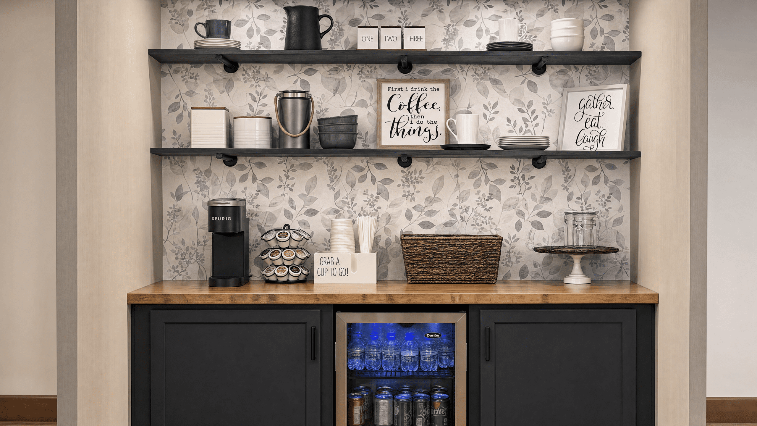 A coffee station with a wooden countertop, black cabinets, and a small refrigerator filled with bottled water. On the countertop, there is a Keurig coffee maker, a cup holder with pods, a box labeled 