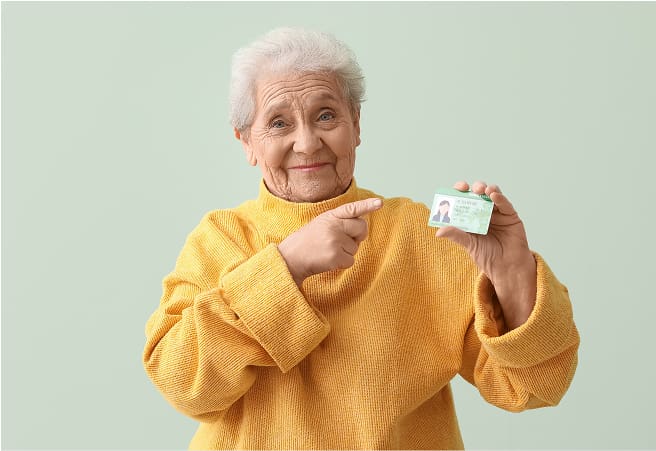 An elderly woman with white hair, wearing a yellow sweater, smiling and pointing at her driver's license.