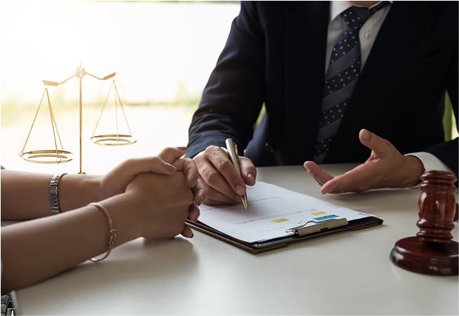 A person in a suit and tie is sitting at a table, holding a pen and signing a document. Another person’s hands are resting on the table, and a wooden gavel is nearby. In the background, there is a scale of justice.