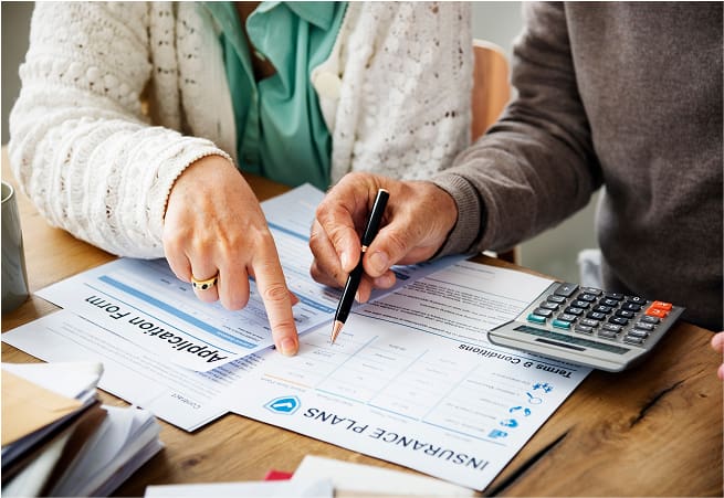 Two people are sitting at a table reviewing insurance documents, with one person pointing and the other holding a pen near a form and a calculator.