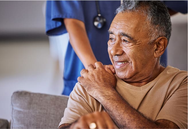 An elderly man sitting on a couch smiling, with a healthcare worker in blue scrubs standing behind him, gently placing a hand on his shoulder.