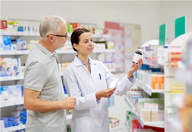 A pharmacist in a white coat shows a medication bottle to an elderly man in a pharmacy aisle filled with various medicine boxes and bottles on the shelves.